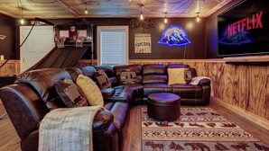 A cozy living room with a sectional sofa, a flat-screen TV, and a neon sign. - Escape Route Retreat · Town Center Retreat* Hot Tub, Game Room, Fire Pit (Lincoln)