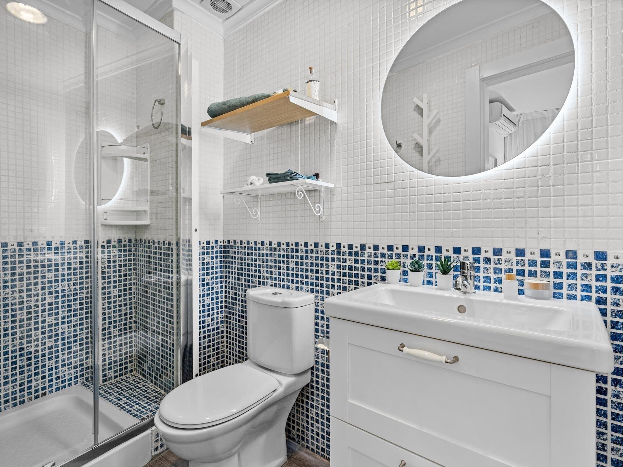 A bathroom with a glass shower enclosure, a toilet, a sink with a mirror, and blue-tiled walls.