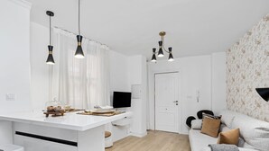 Une cuisine moderne avec des meubles blancs, un coin repas avec une table et des chaises blanches, et un salon avec un canapé gris et un papier peint à motifs.