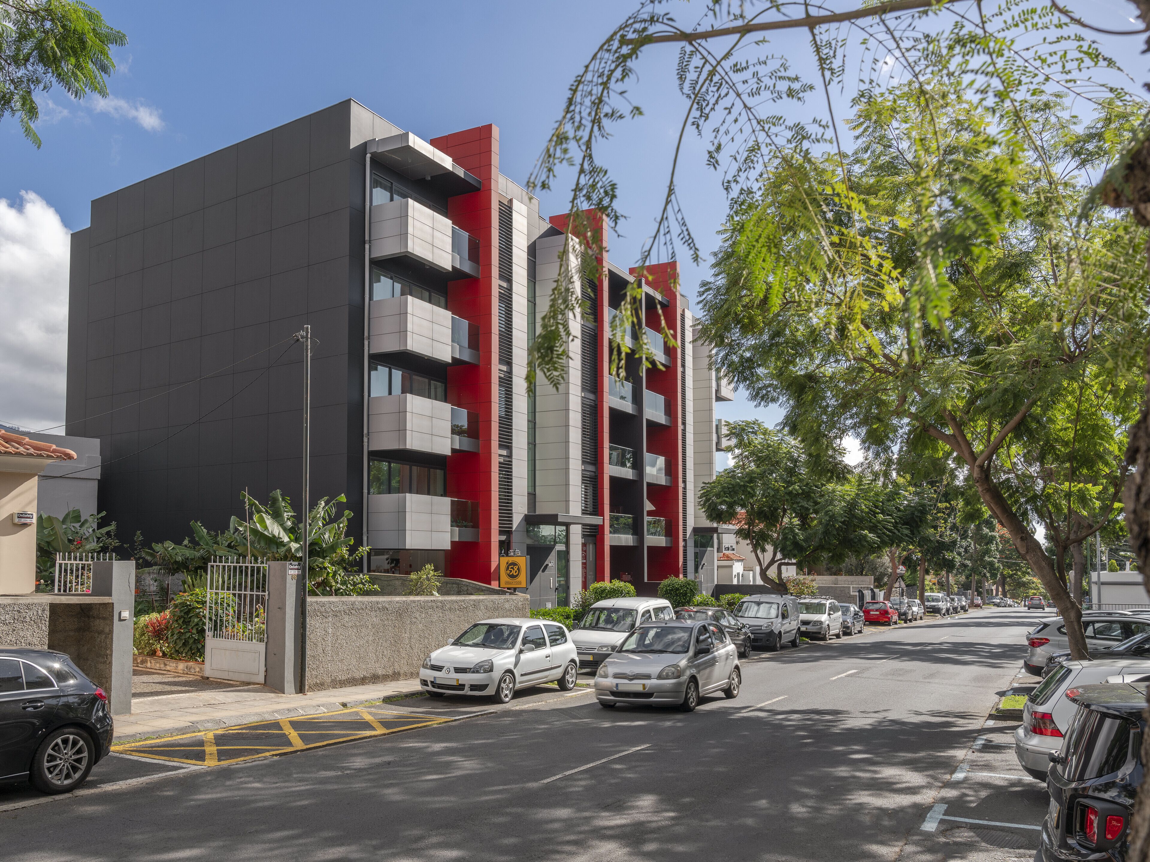 A modern building with multiple balconies, a parking lot with cars, and surrounding greenery.