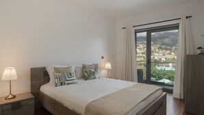 A bedroom with a large bed, two bedside lamps, a view of a city through a large window, and a wooden nightstand. - City Holidays in Madeira - Madalenas XXI (Funchal)