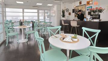 A restaurant with teal chairs and white tables, a receptionist at the counter, and a view of the sea through large windows.