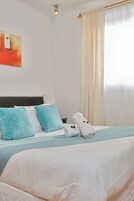 Una cama bien hecha con ropa de cama blanca y aguamarina, un par de toallas enrolladas y una manta aguamarina.