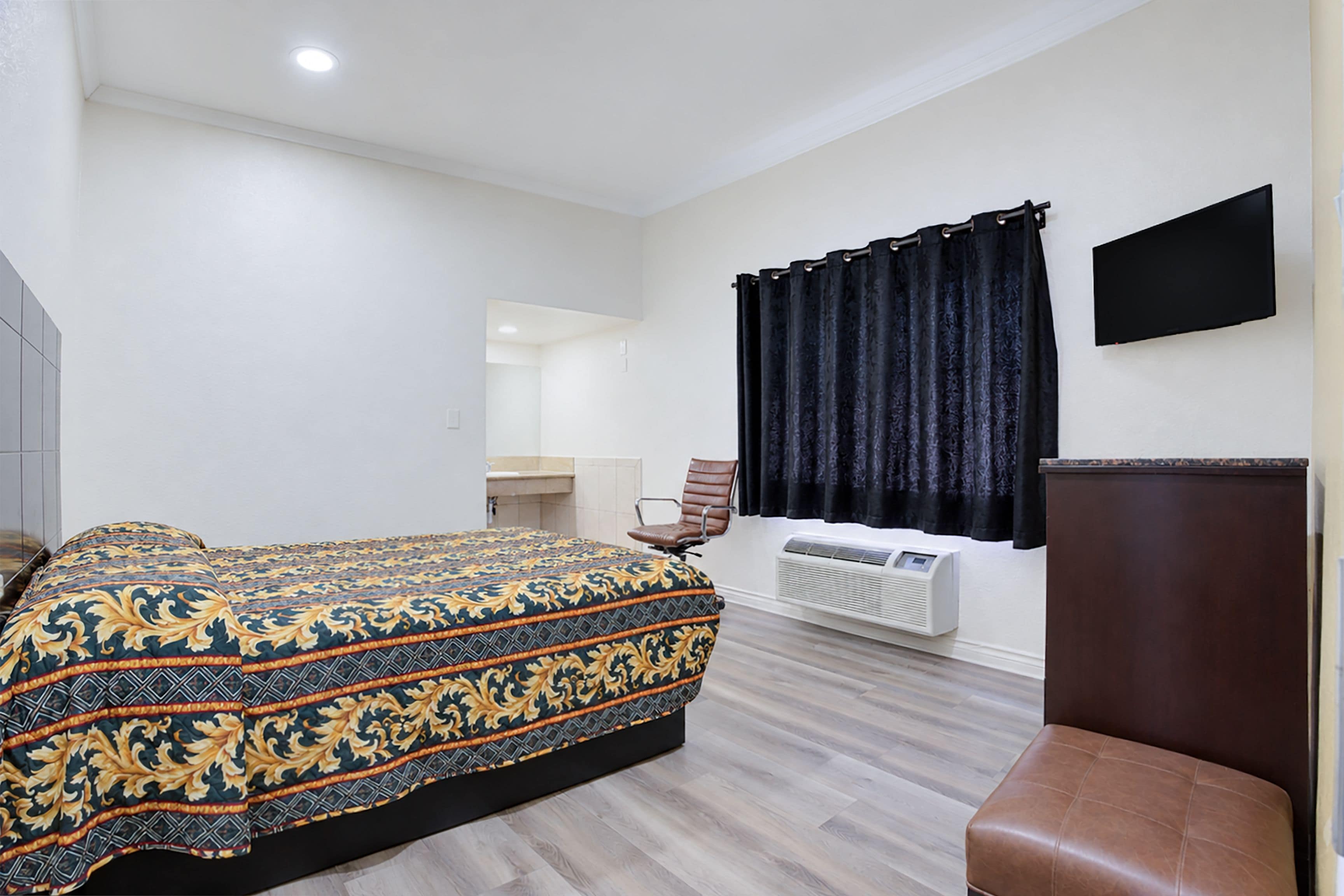 A hotel room with a bed, a chair, a flat-screen TV, a window with curtains, and a wall-mounted air conditioner.