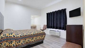 A hotel room with a bed, a chair, a flat-screen TV, a window with curtains, and a wall-mounted air conditioner.