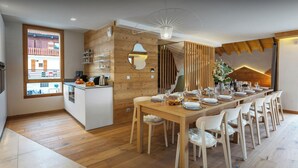 A modern kitchen with a wooden island, a dining area with a long table and chairs, and a view of a building through a window. - Chalet Mevi - family break in the Alps 13p, great location - OVO Network (Crest-Voland)