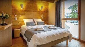 A modern bedroom with a wooden headboard, a large bed with white bedding, two wall-mounted lamps, and a view of a snowy landscape through a window. - Chalet Mevi - family break in the Alps 13p, great location - OVO Network (Crest-Voland)