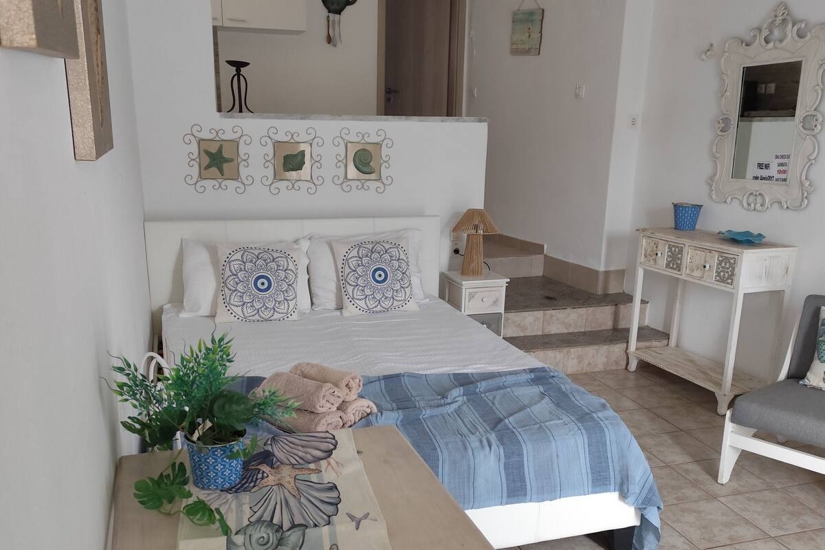 Un dormitorio con cama, mesita de noche y tocador. Hay una ventana con cortinas, un espejo en la pared y una puerta que da a otra habitación.