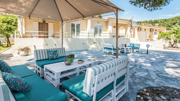 An outdoor seating area with white furniture and green cushions, a covered patio, and a stone floor. - Alexia studios (Skiathos)