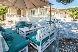 Un espace extérieur aménagé avec des meubles blancs et des coussins verts, une terrasse couverte et un sol en pierre.