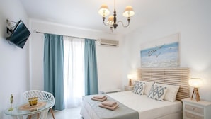 A bedroom with a bed, bedside lamps, a TV, a chair, and a window with curtains. - Alexia studios (Skiathos)