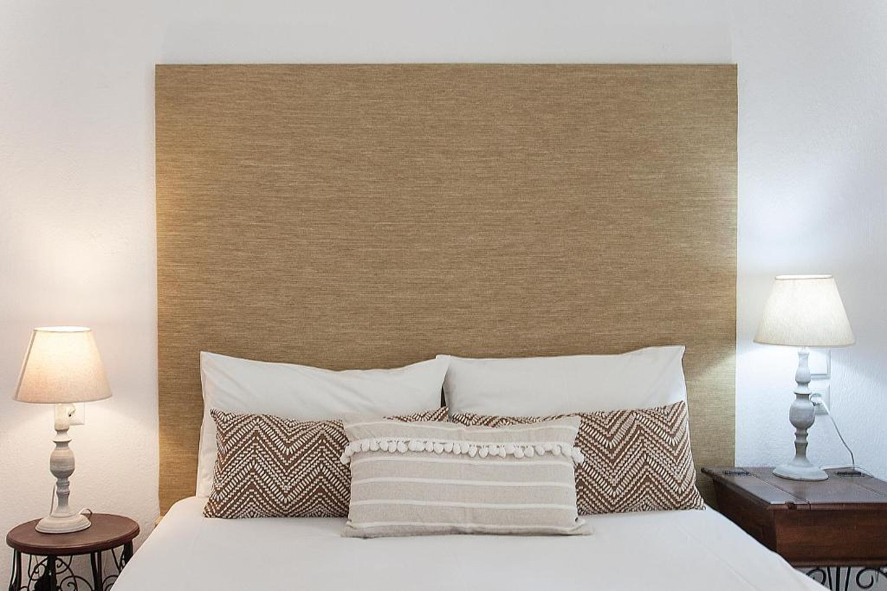 Un lit bien fait, avec du linge de lit blanc et des coussins à motifs, flanqué de deux tables de chevet sur lesquelles sont posées des lampes.