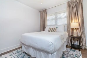 Un dormitorio ordenado con cama, mesita de noche, lámpara y cortinas.
