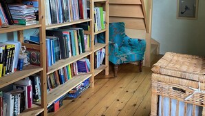 A room with wooden bookshelves filled with books, a blue chair, a wicker basket, and a staircase. - Comfortable house in the heart of Normandy (Flancourt-Crescy-en-Roumois)