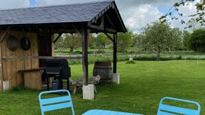 A gazebo with a grill, two blue chairs, and a wooden building with a wheel in the background. - Comfortable house in the heart of Normandy (Flancourt-Crescy-en-Roumois)
