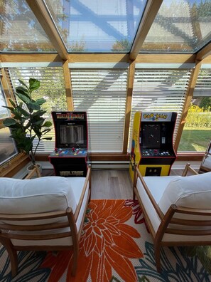 Relax in a spacious room with a large window offering a view of the outdoors, featuring arcade games, comfortable seating, and a touch of greenery.