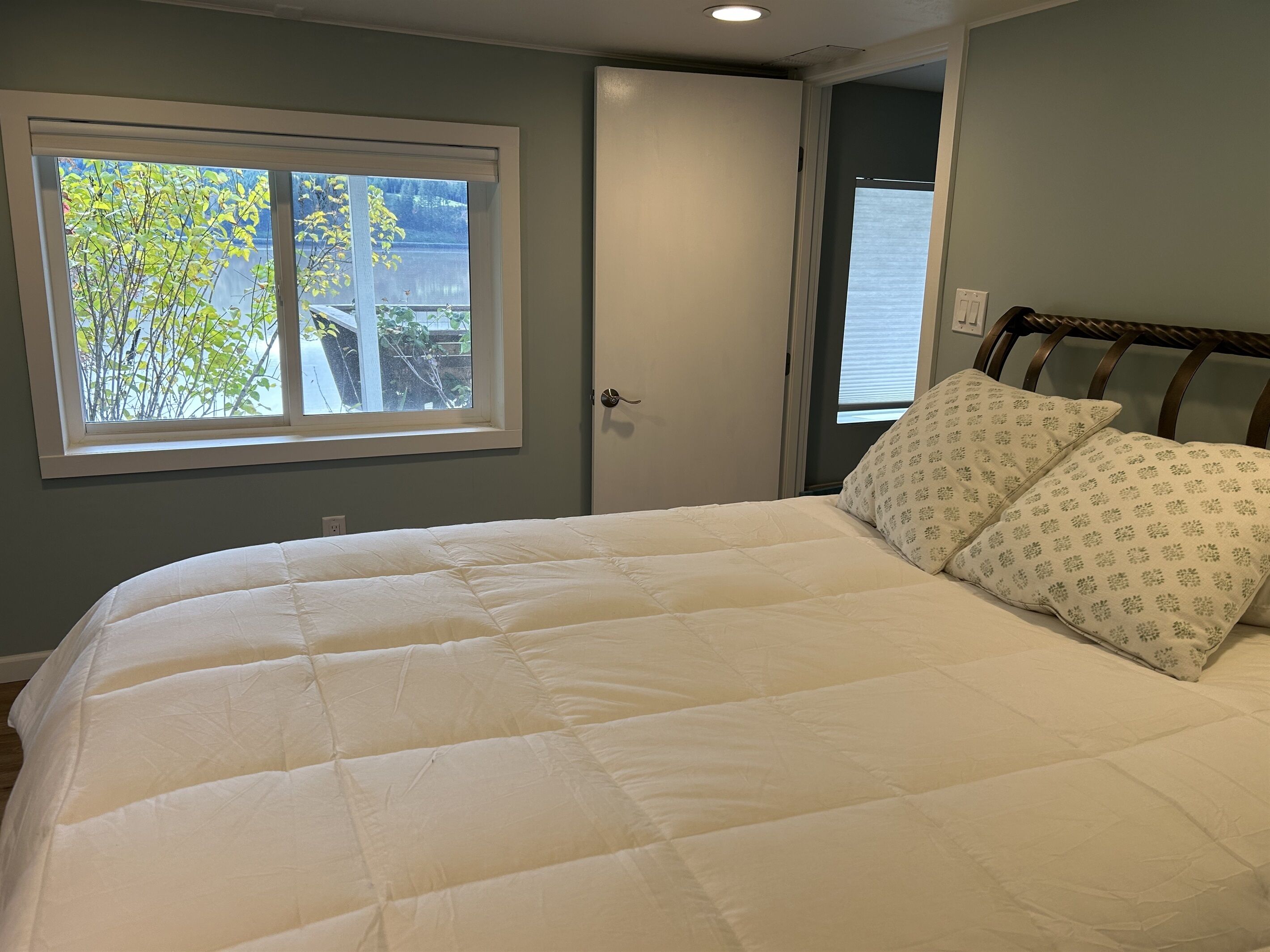 A bedroom with a large bed, a window with a view of trees, and a door leading to another room.