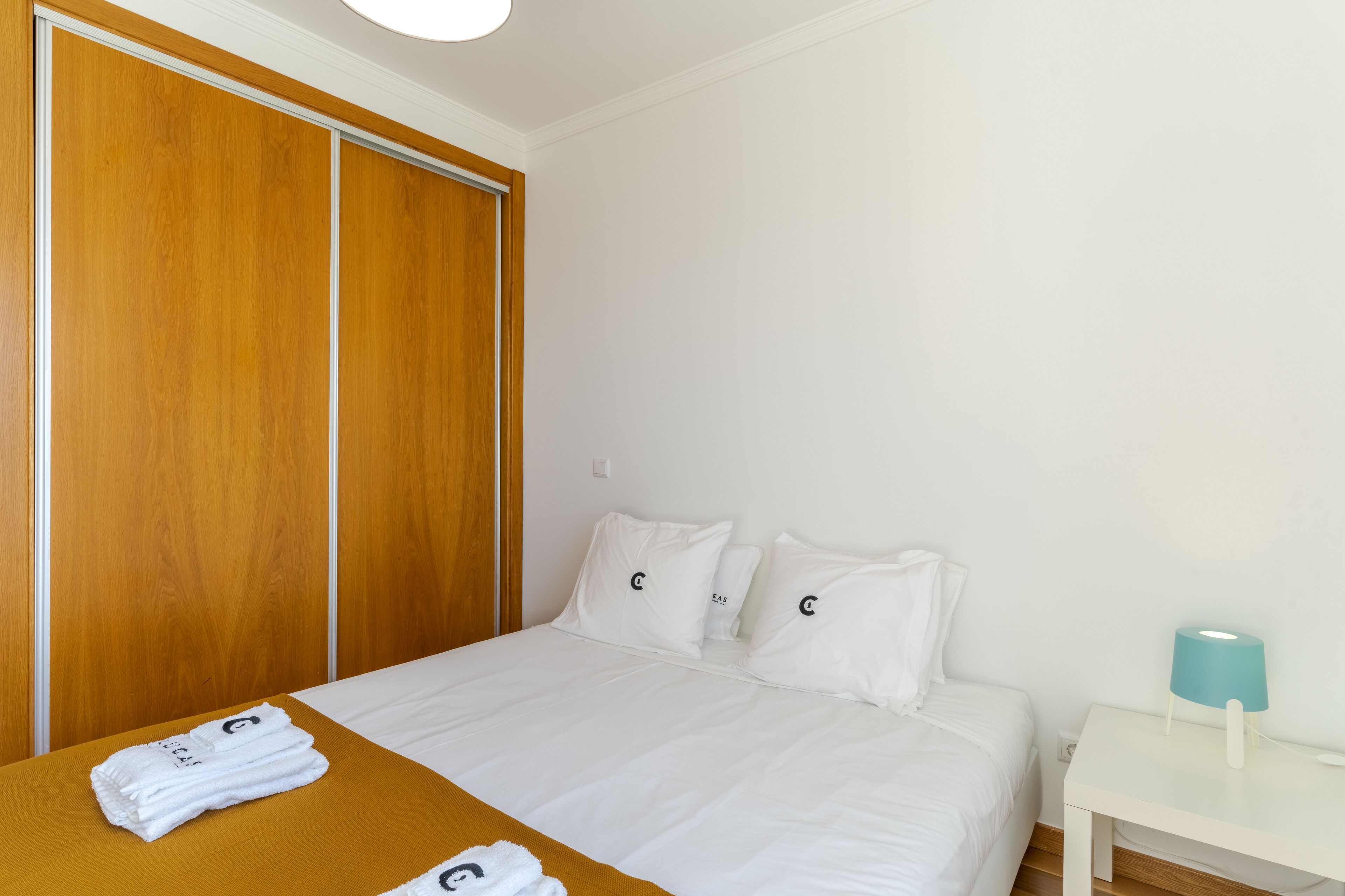 A hotel room with a bed, white bedding, a wooden wardrobe, a white nightstand with a lamp, and towels with a logo.