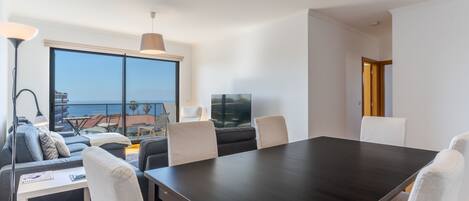 A modern living room with a dark wooden dining table, white chairs, a sofa, and a large window offering a view of the sea.