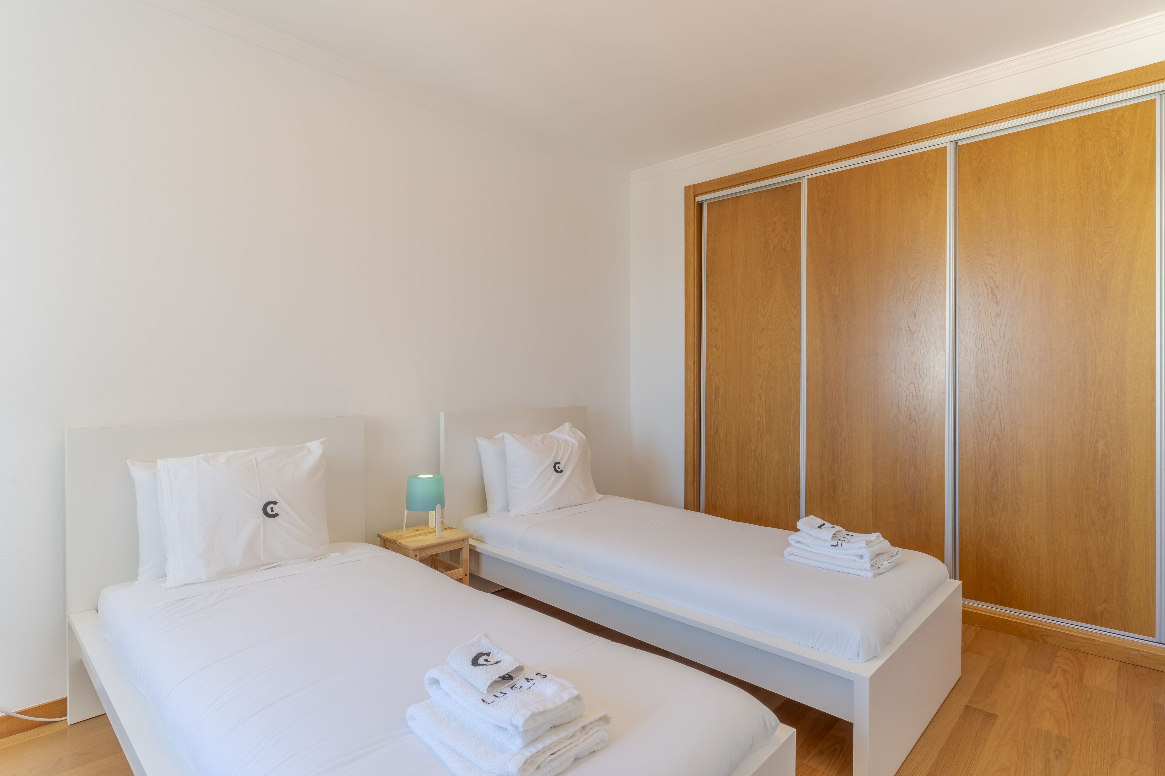 A hotel room with two single beds, a wooden wardrobe, a bedside table with a lamp, and neatly folded towels.