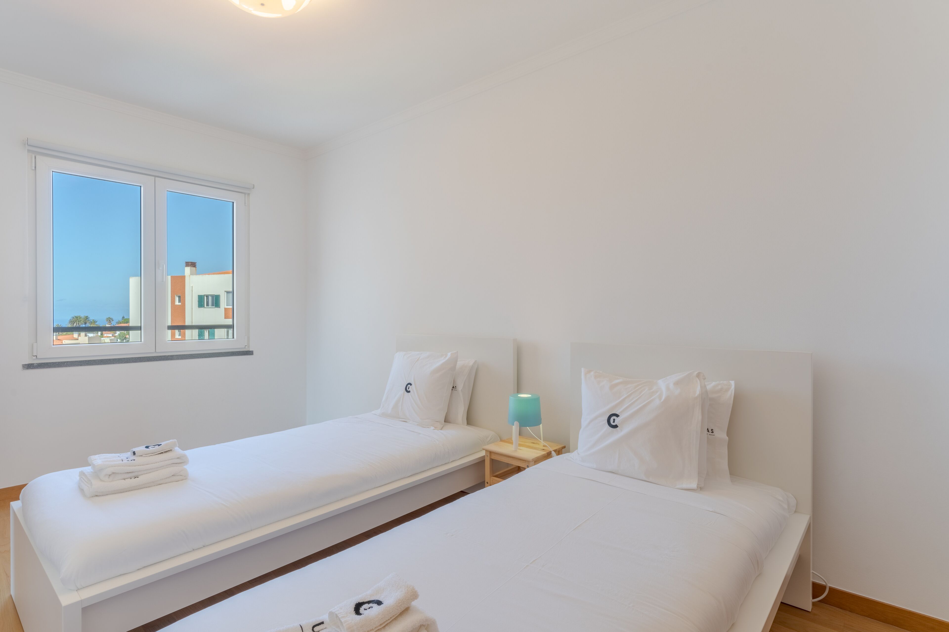 Ett hotellrum med två separata sängar, ett litet bord med en lampa, ett fönster med utsikt över byggnader och en välordnad säng med handdukar.