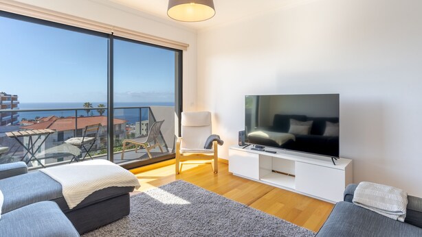 A modern living room with a large TV, comfortable seating, and a balcony with a view of the sea. - Jardins do Oceano (Santa Cruz)