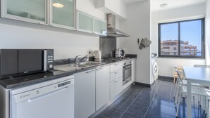 A modern kitchen with a dishwasher, microwave, sink, and stove. There is a window with a view of buildings outside. - Jardins do Oceano (Santa Cruz)