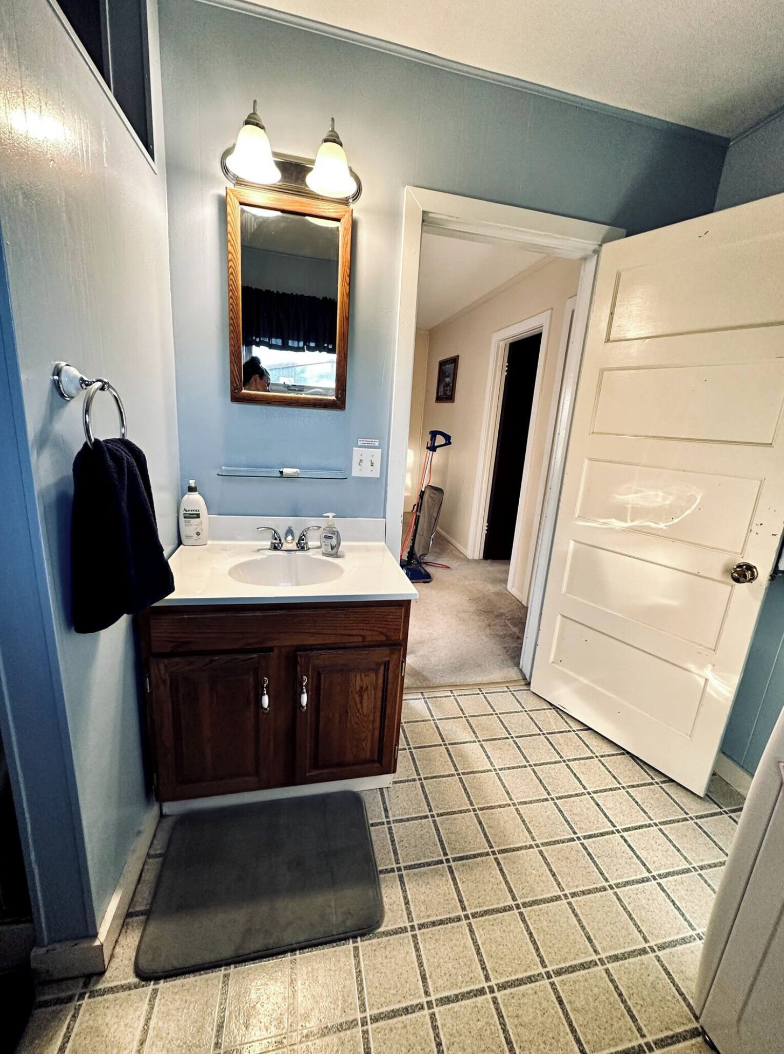 A bathroom with a white sink, wooden vanity, two lights above the mirror, and a door leading to another room.