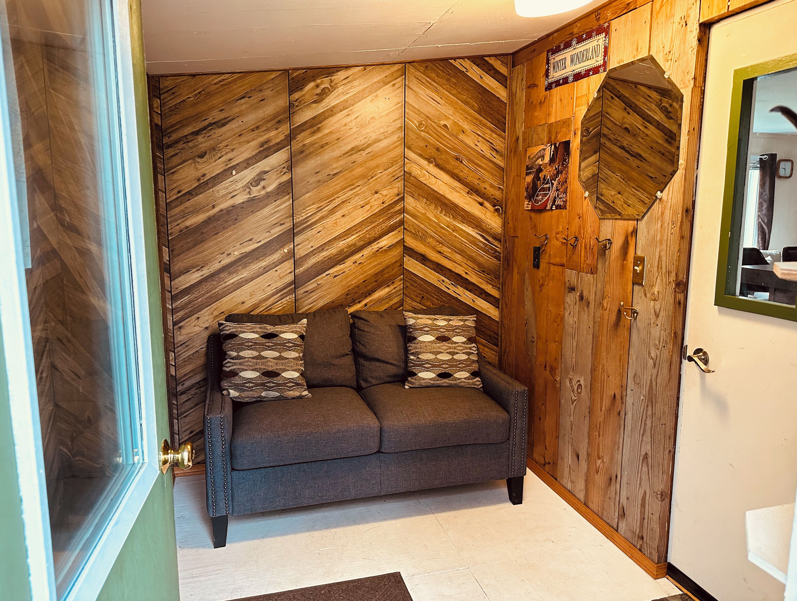 Un cuarto acogedor con un muro de madera con diseño de espiga, un sofá gris con cojines estampados y una puerta de madera con un cartel encima.