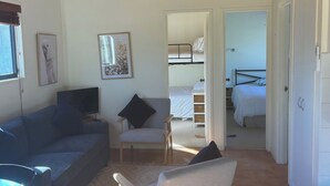 A spacious room with a high wooden ceiling, a sofa, a chair, a TV, and a view into a bedroom with a bunk bed. - River Front Cabin (Bridgewater on Loddon)
