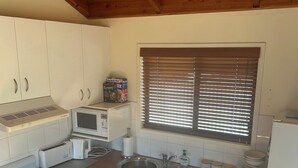 A compact kitchen with white cabinets, a stove, microwave, sink, and a window with blinds. - River Front Cabin (Bridgewater on Loddon)