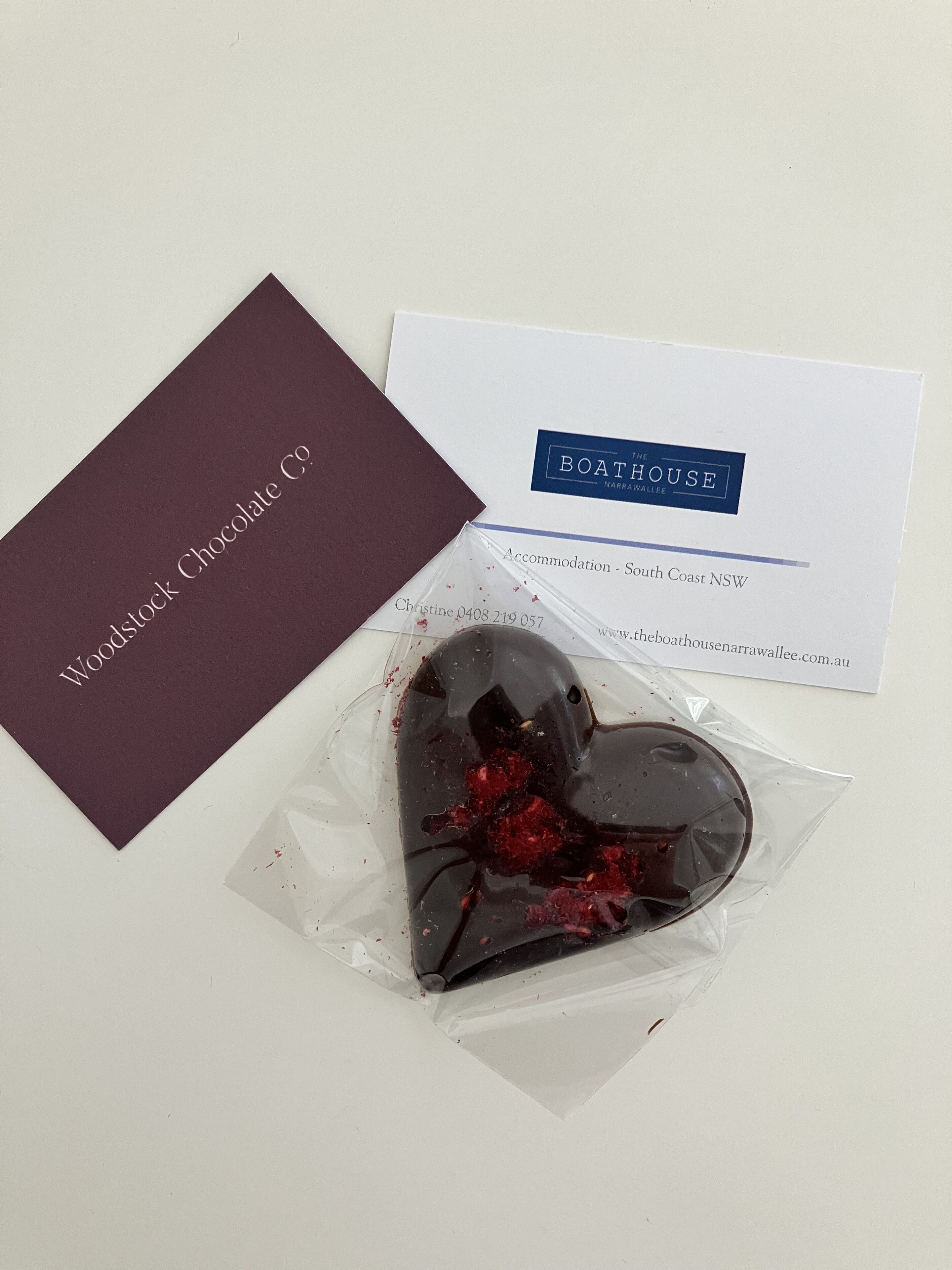 Une carte de visite, une carte de couleur bordeaux et un chocolat en forme de cœur avec un fourrage rouge.