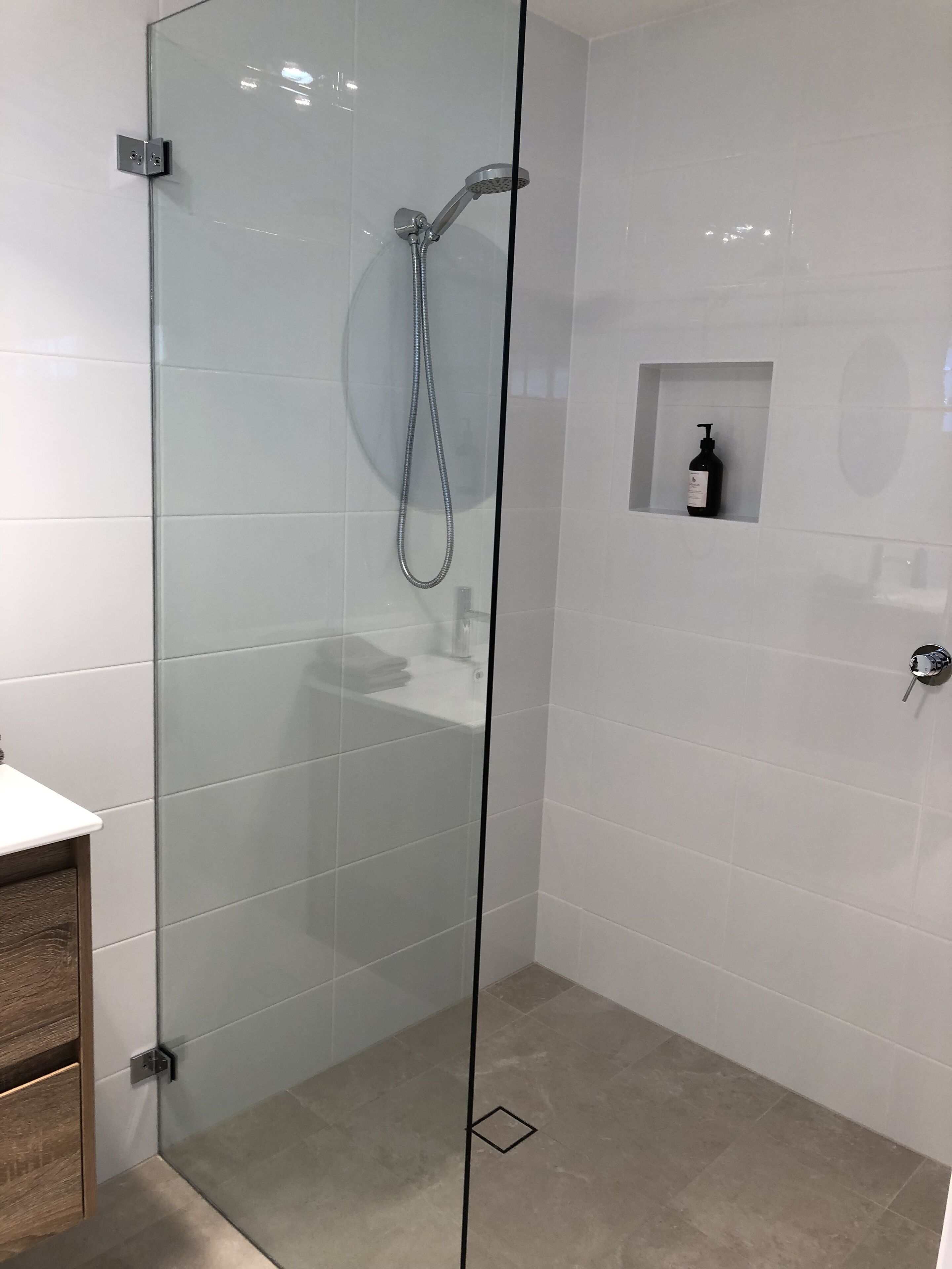 Cabina de ducha con paredes de vidrio, un cabezal de ducha manual, un pequeño estante con una botella y un armario de madera.