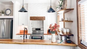 Une cuisine moderne avec des meubles blancs, des appareils en acier inoxydable, des plans de travail en bois, des luminaires suspendus et un mur de fond en briques.