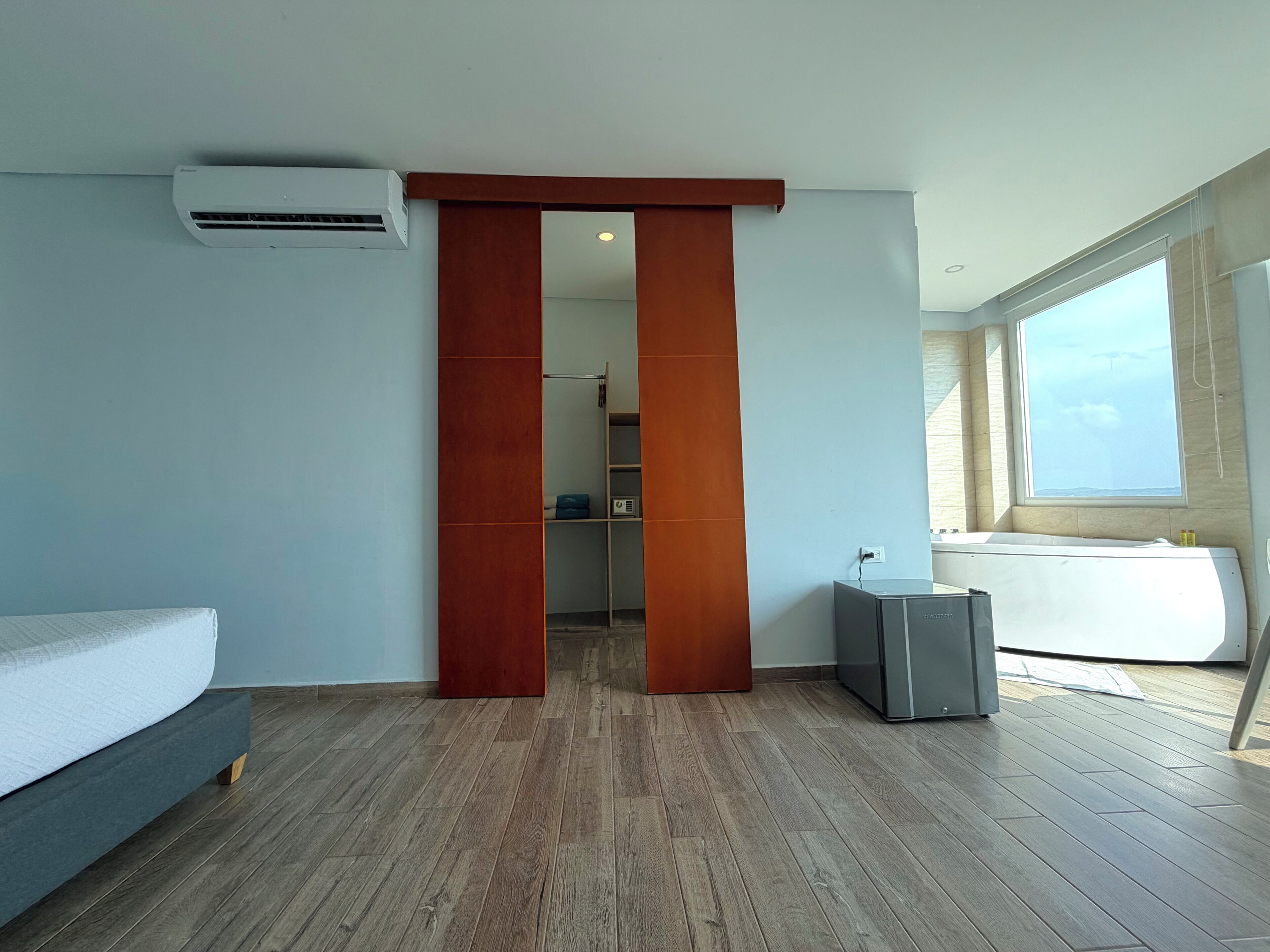 A modern hotel room with a wooden floor, a bed, a desk, an air conditioning unit, and a window with a view.