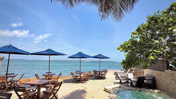 Une terrasse au bord de la plage, avec des tables en bois et des parasols bleus, une piscine et une vue sur la mer.