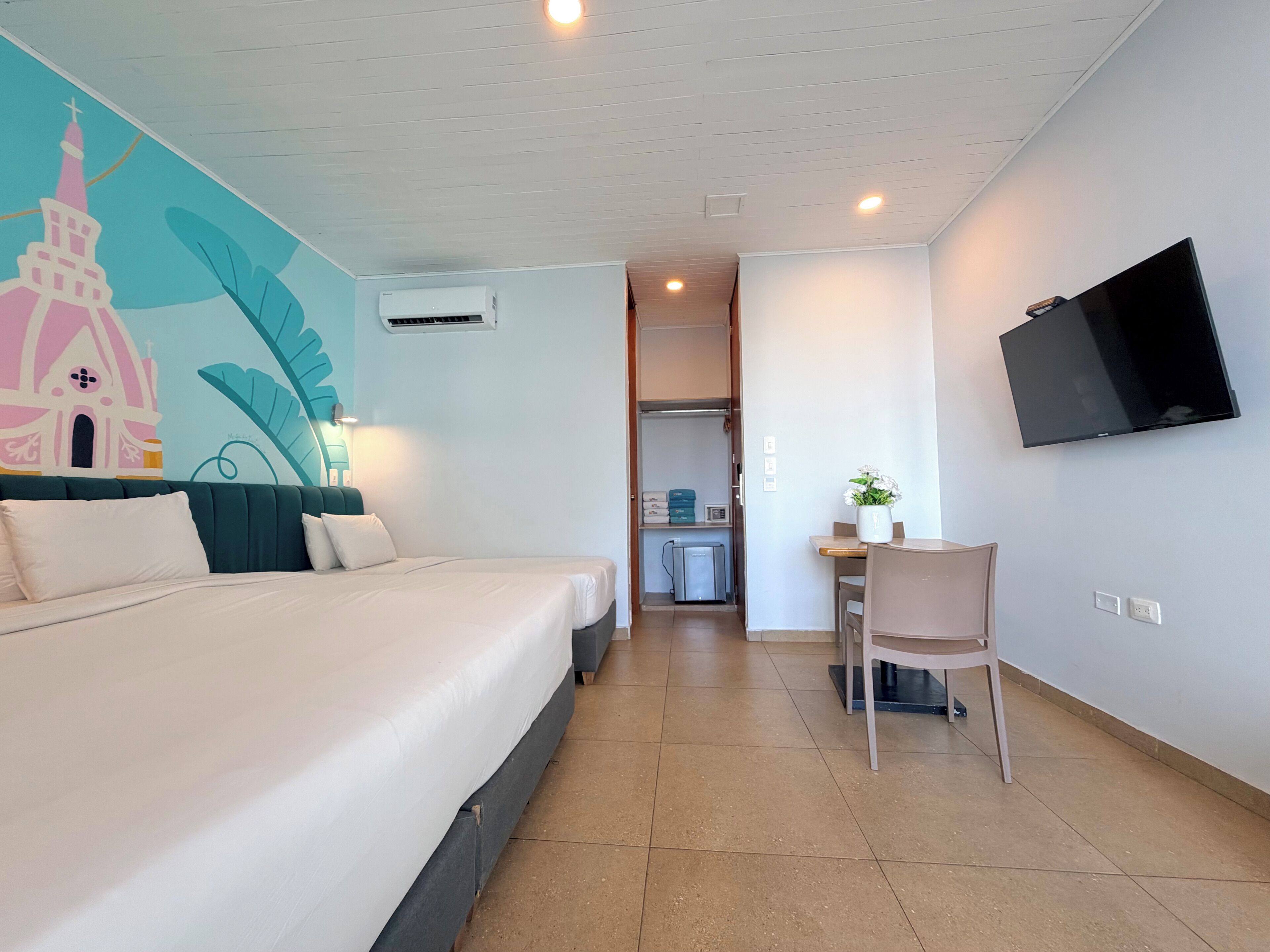 A hotel room with a bed, a desk with a chair, a TV mounted on the wall, and a mural on the wall.