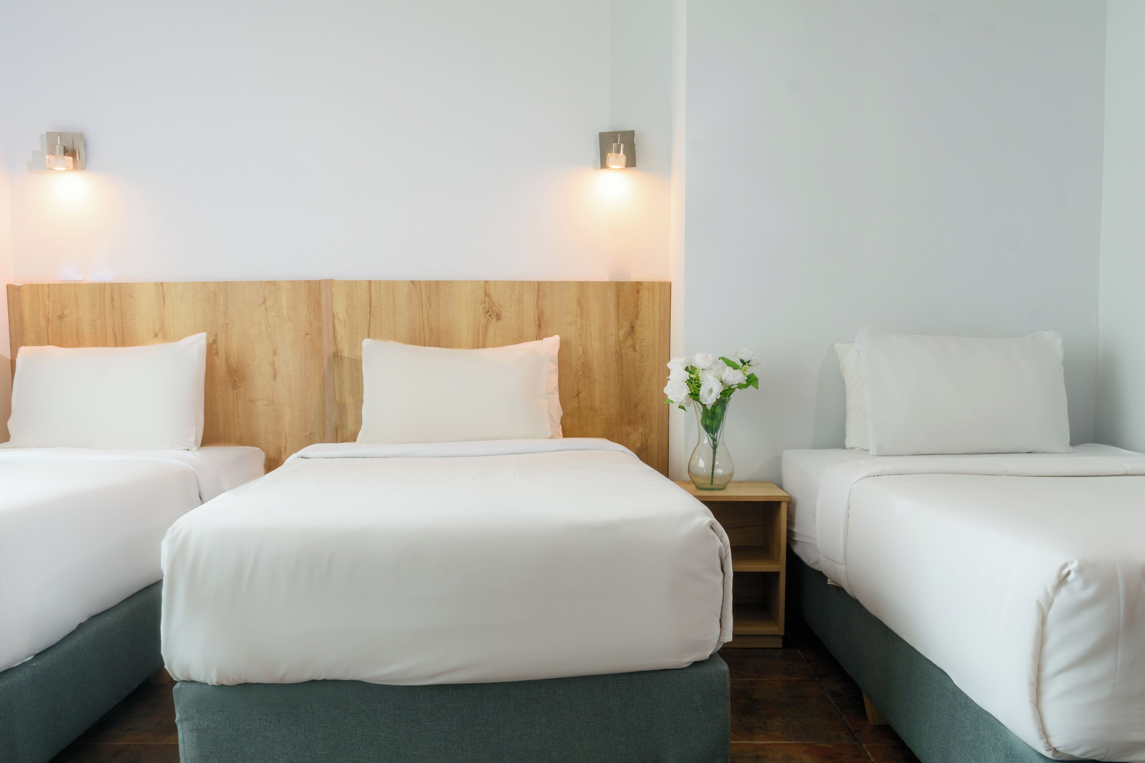 Una habitación de hotel con tres camas individuales, cada una con ropa blanca y cabeceros de madera. Hay dos lámparas de pared a cada lado del cabecero.