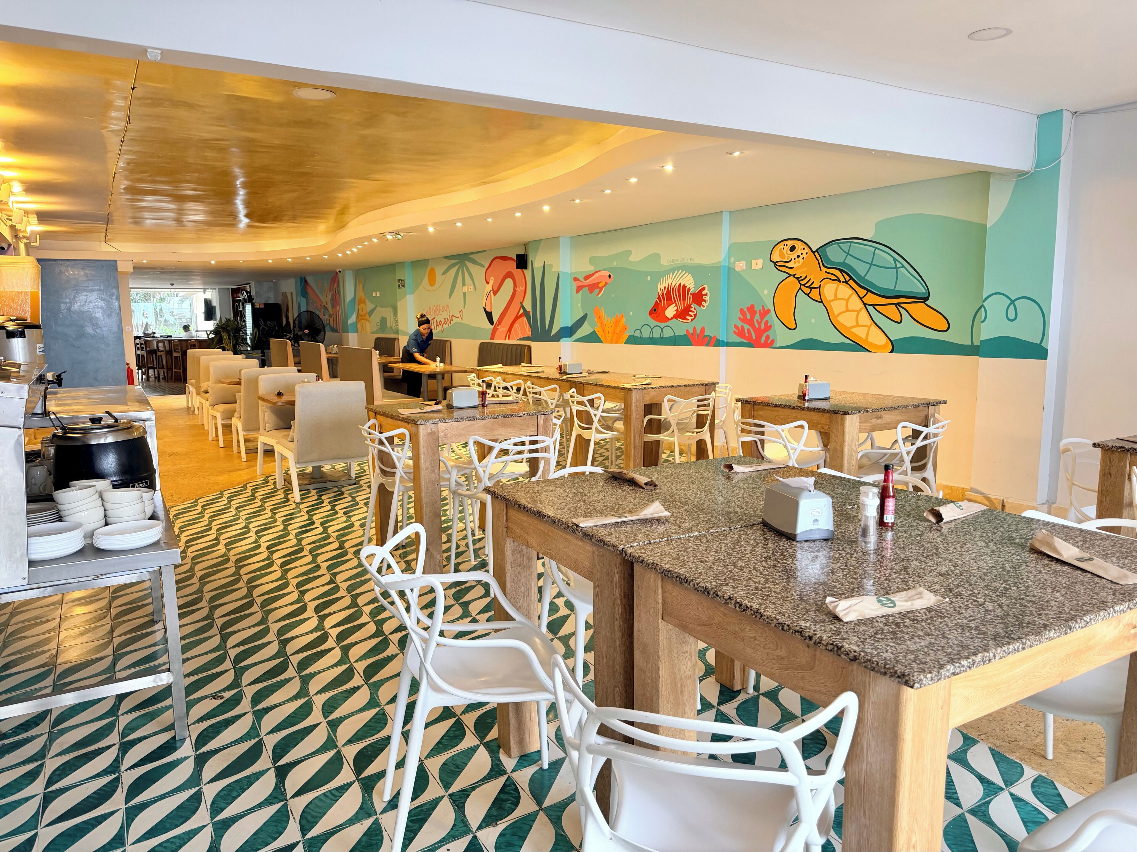 Un restaurant décoré d’une fresque murale aux couleurs vives, avec des chaises blanches et un sol à motifs.