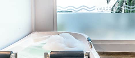 A bathtub with a view of palm trees and a body of water through a frosted glass window.