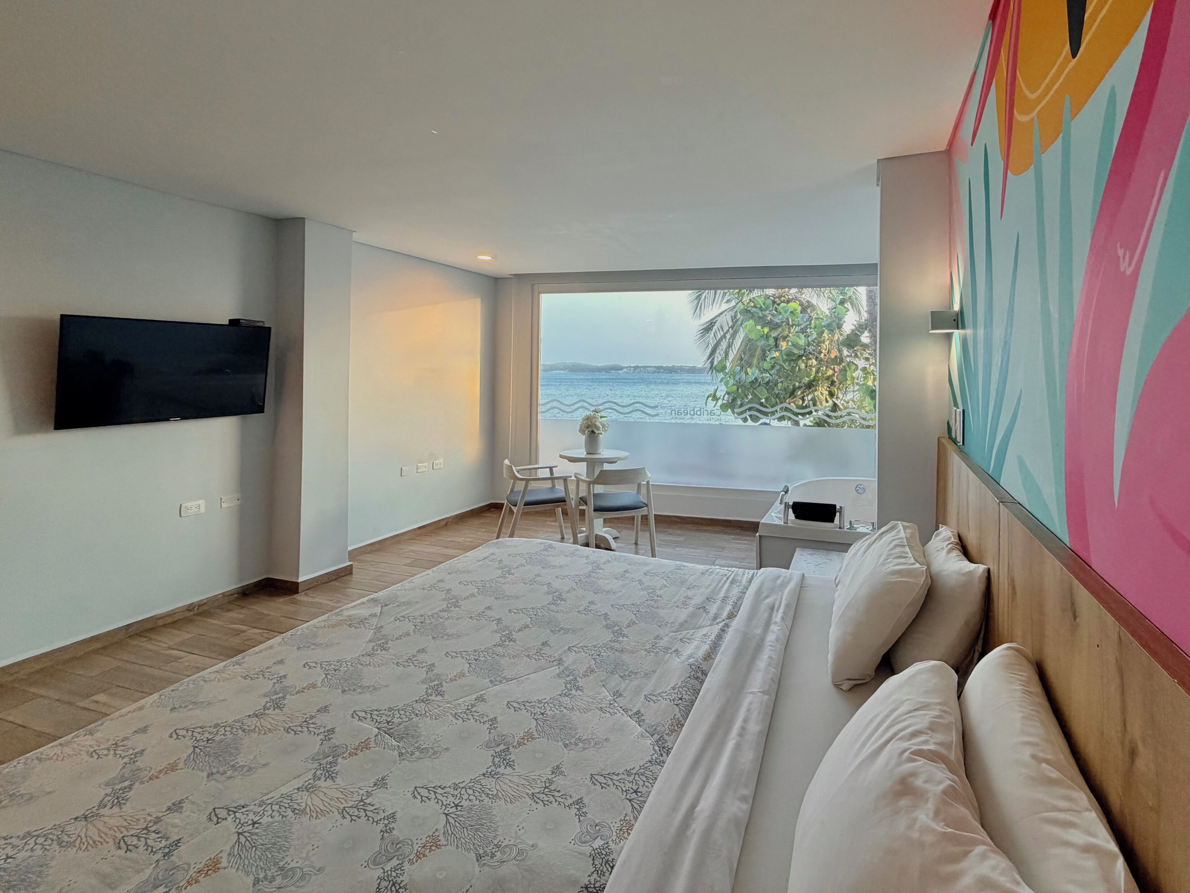A bedroom with a large bed, a TV mounted on the wall, a small table with chairs, and a view of the sea.