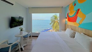 Une chambre d’hôtel avec un lit, une télévision, une petite table avec une chaise, une fenêtre donnant sur la mer et une fresque colorée sur le mur.