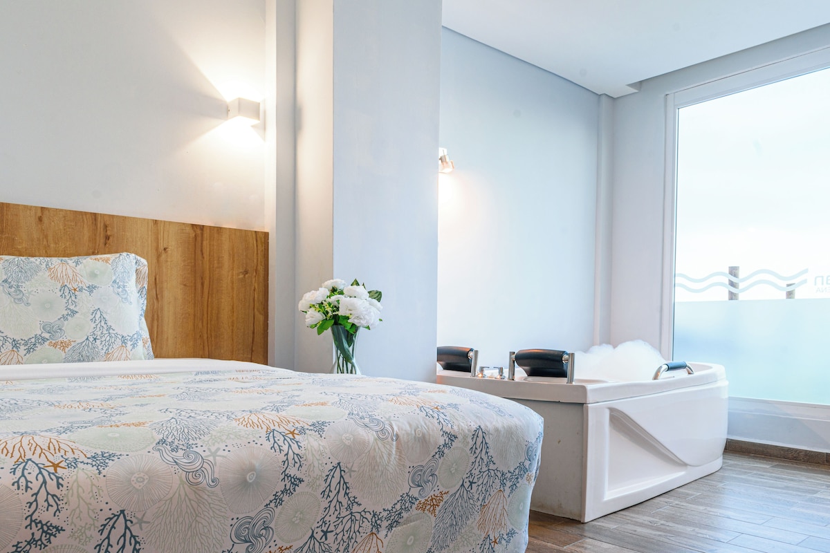 A modern bedroom with a wooden headboard, a large bed with a patterned bedspread, a bedside table with a vase of flowers, a built-in bathtub, and a large window with a view.