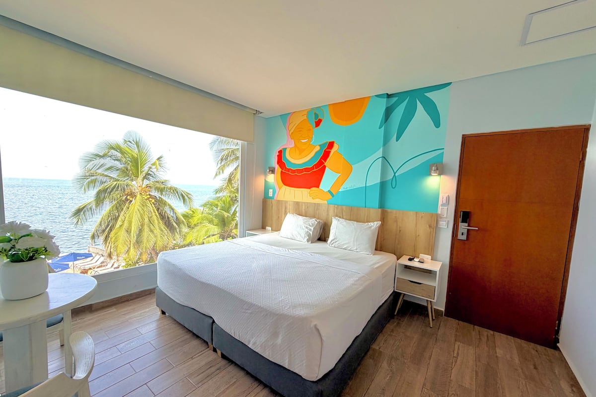Ein Hotelzimmer mit einem Bett, Nachttischen, einem Schreibtisch mit Stuhl, einem großen Fenster mit Blick auf das Meer und Palmen sowie einem farbenfrohen Wandgemälde.
