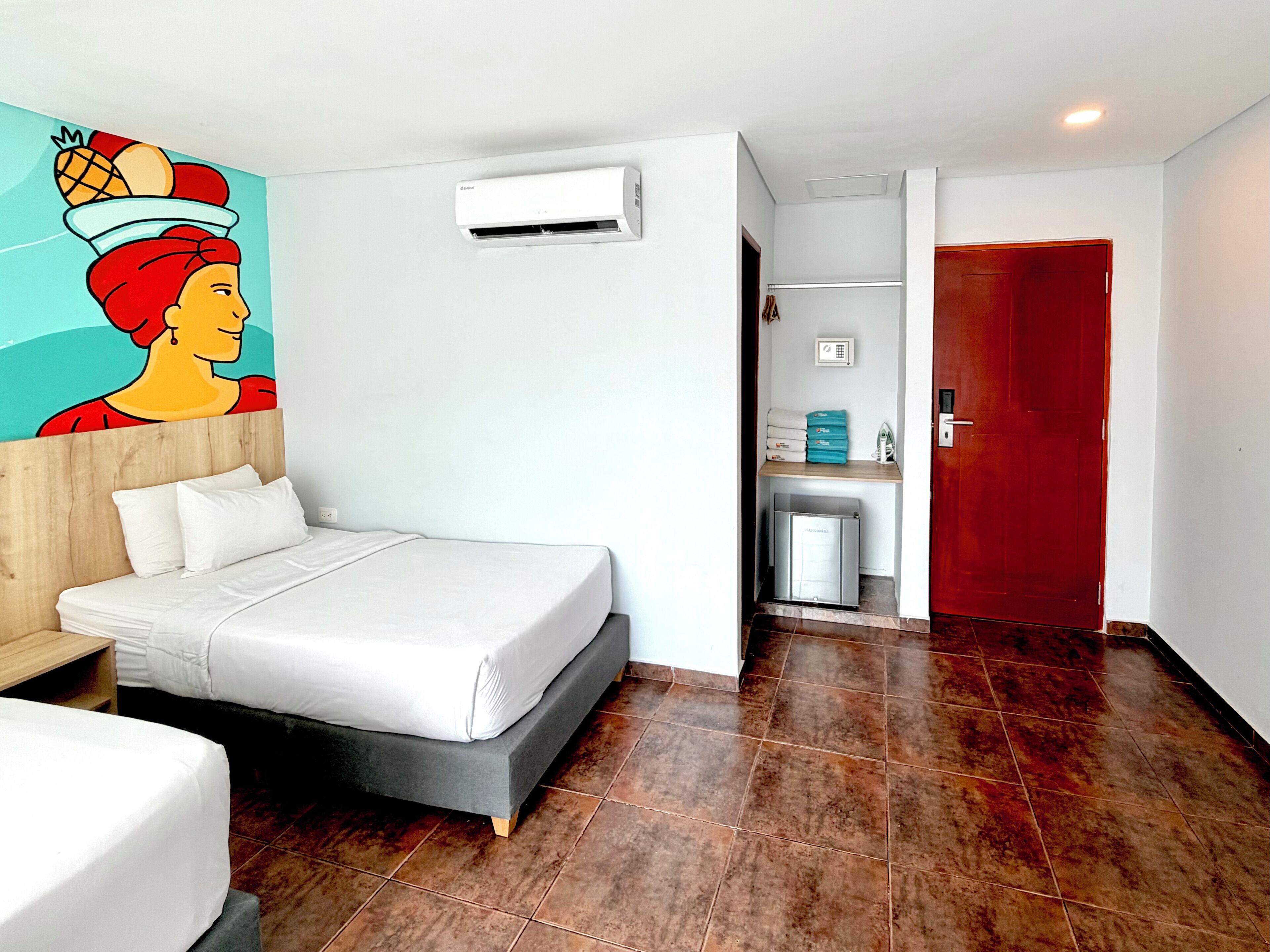 Habitació d'hotel amb llit, aire condicionat fixat a la paret, porta vermella i zona de bany petita.