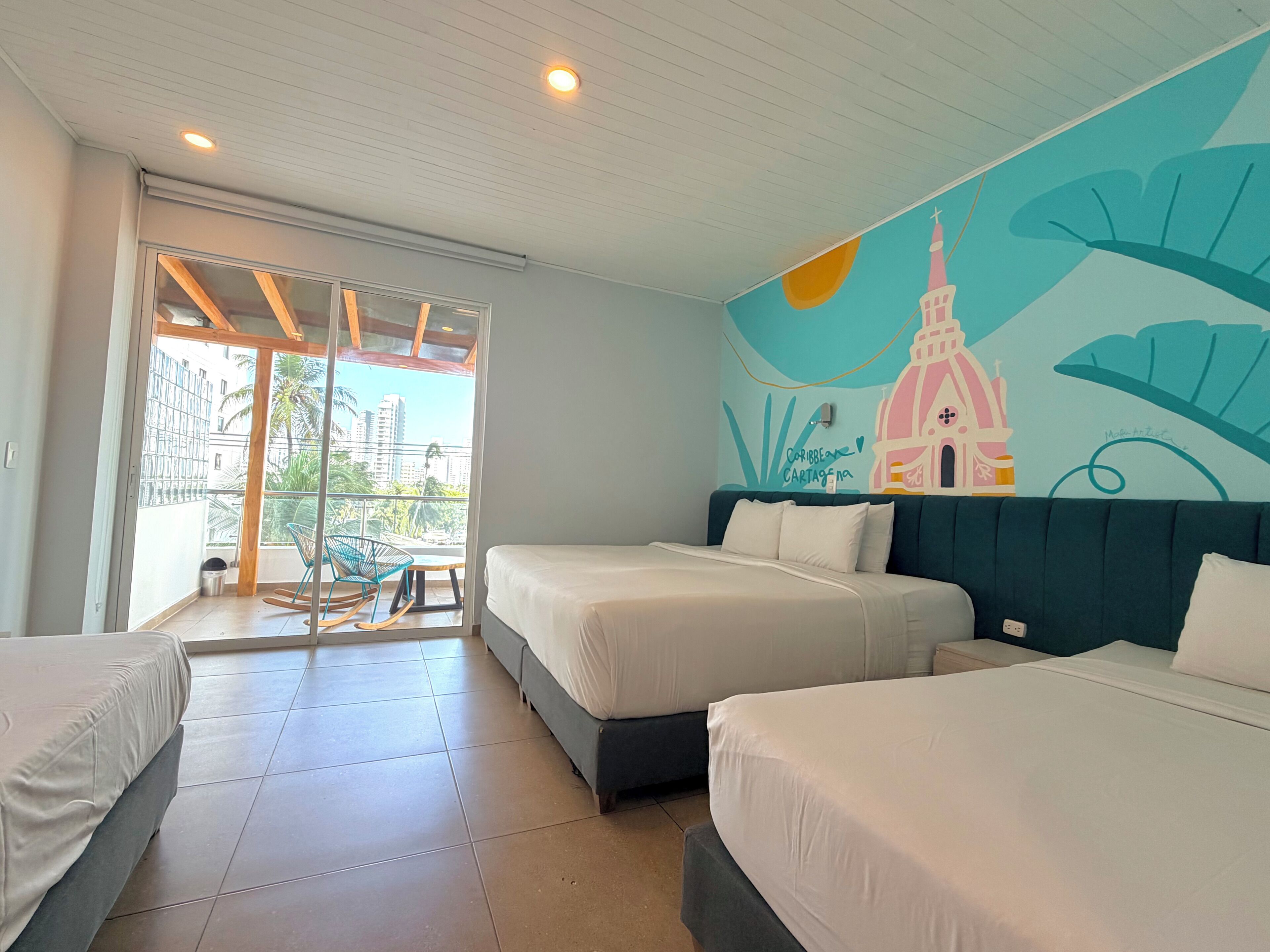 Hotelkamer met twee bedden, een balkon met uitzicht op palmbomen en gebouwen, en een muurschildering.