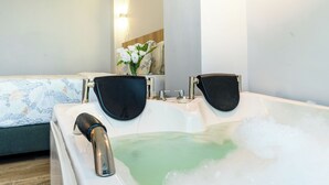 Une salle de bain avec une baignoire, deux pommes de douche noires, un arrangement floral et un lit recouvert d’une housse à motifs.