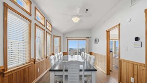 Un'ampia sala da pranzo con un lungo tavolo e sedie bianche, pareti rivestite in legno e grandi finestre con tende.