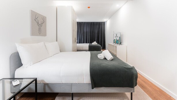 A modern hotel room with a bed, a glass table, a grey sofa, a black throw blanket, and a framed artwork on the wall. - GuestReady - Uptown suites 1 (Porto)