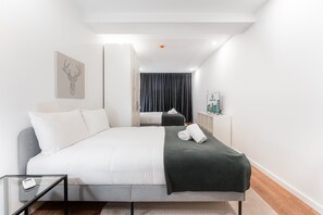 Quarto de hotel moderno com uma cama, uma mesa de vidro, um sofá cinza, um cobertor preto e uma obra de arte emoldurada na parede.